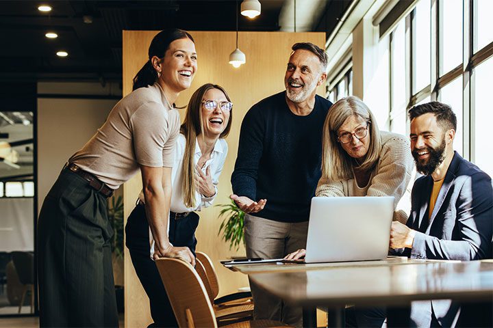 payroll A group of five adults in a modern office laugh and discuss something around a laptop on a table. Large windows let in natural light.